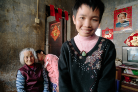 Yongyi Ou, a 12-year-old dysgnosia girl is kissing her grandmother Mei Lin while Yueyi Ou, the older sister also with dysgnosia, is standing in the room and smiling. This is a family with 5 people and 4 of them have mental disease, the 83-year-old lady Mei Lin has to support the family by herself. Yuyang Liu.